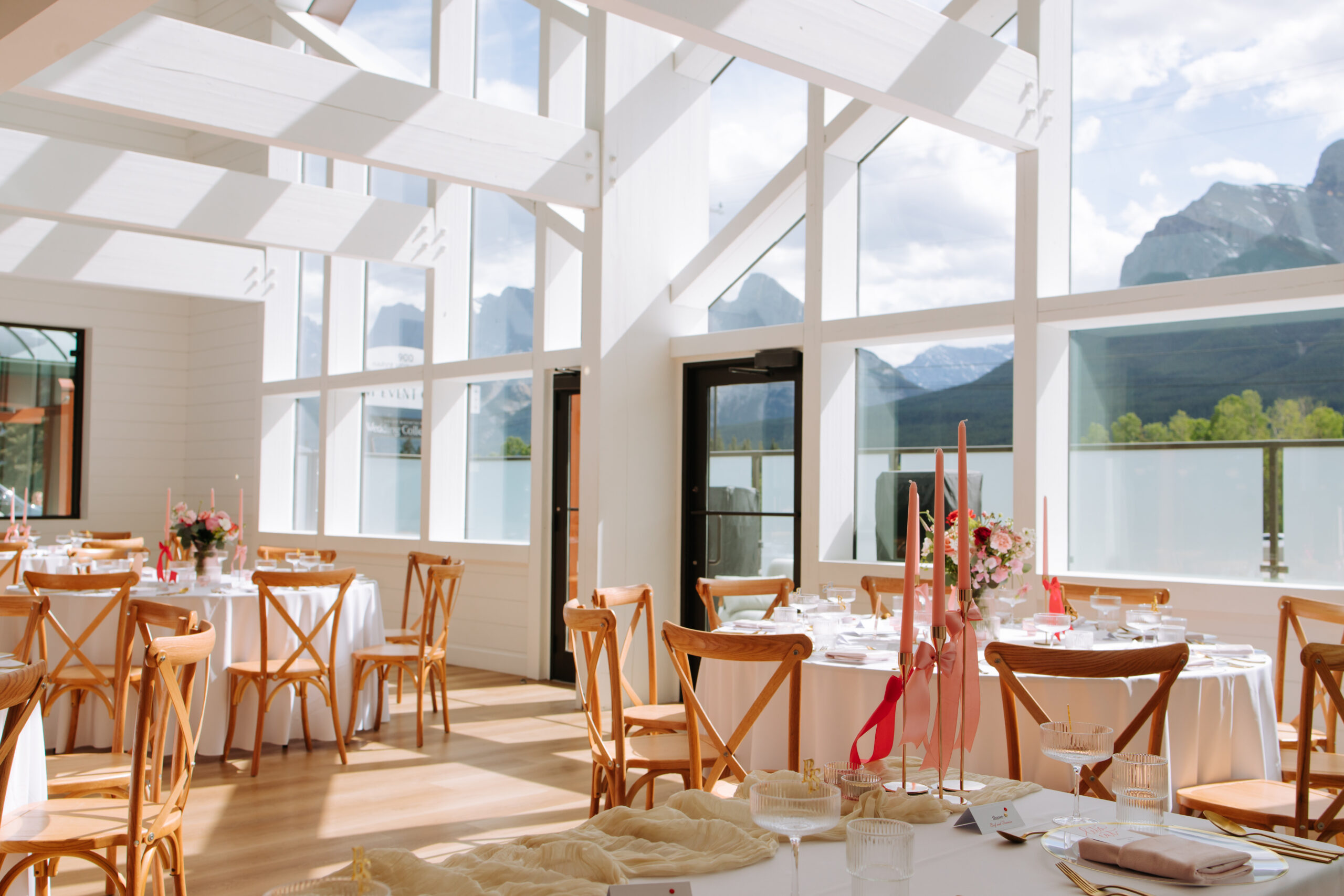 Sunlit wedding reception setup with wooden chairs and pink florals inside The Gem in Canmore, Alberta.
