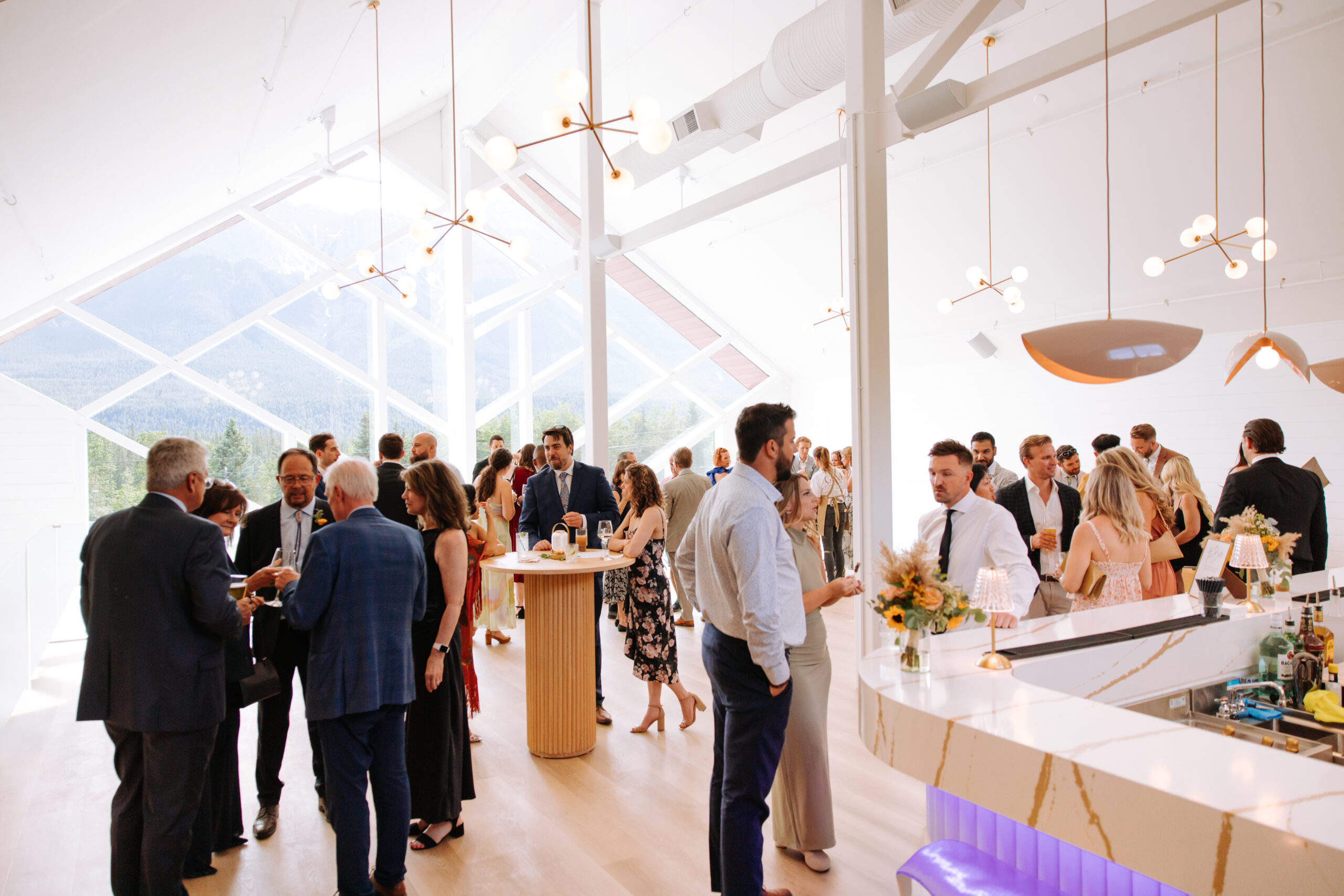 Wedding guests mingle during cocktail hour inside The Gem, with floor-to-ceiling windows showcasing mountain views.