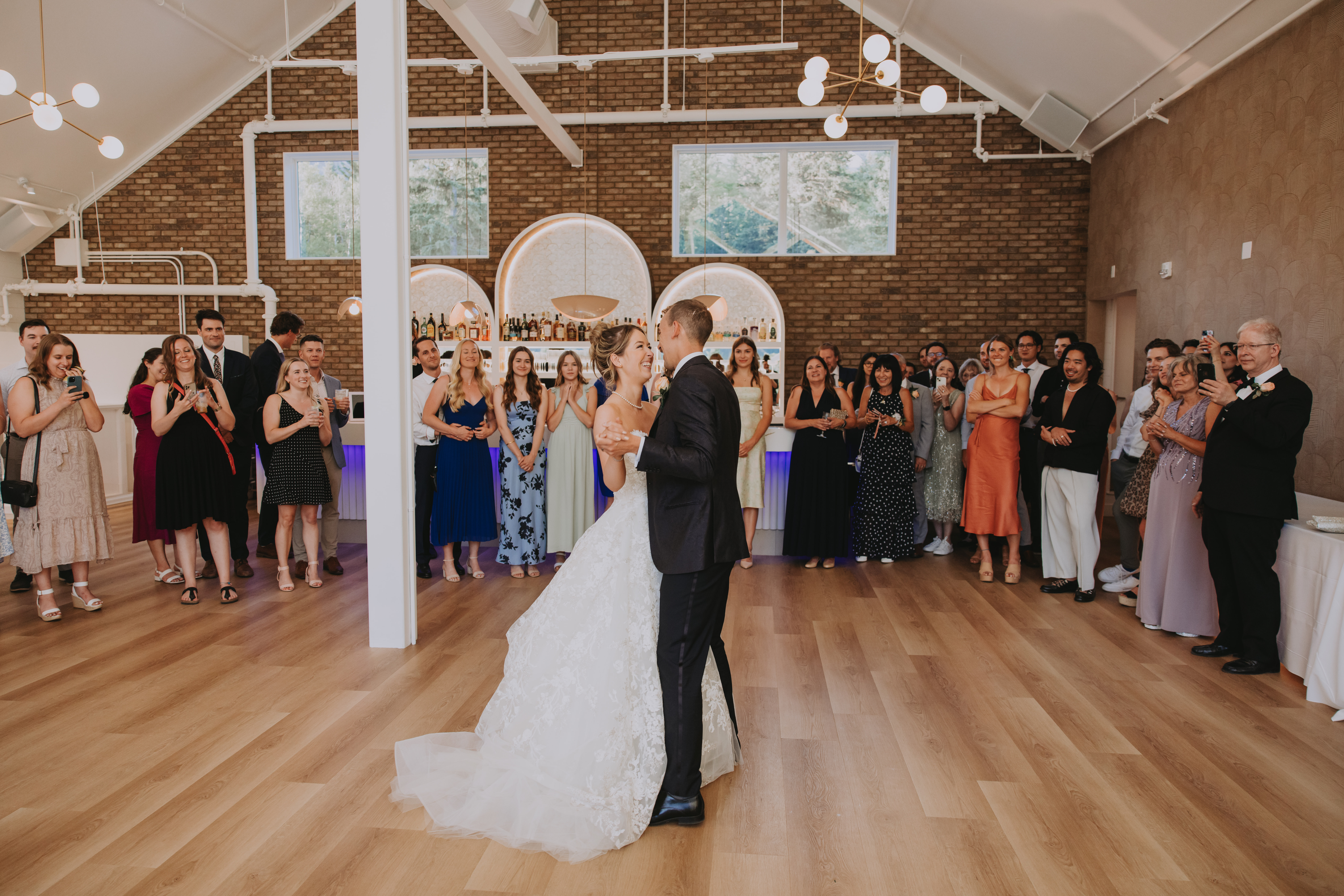 Bride and groom sharing their first dance inside The Gem venue in Canmore, with guests gathered around and a brick bar backdrop.