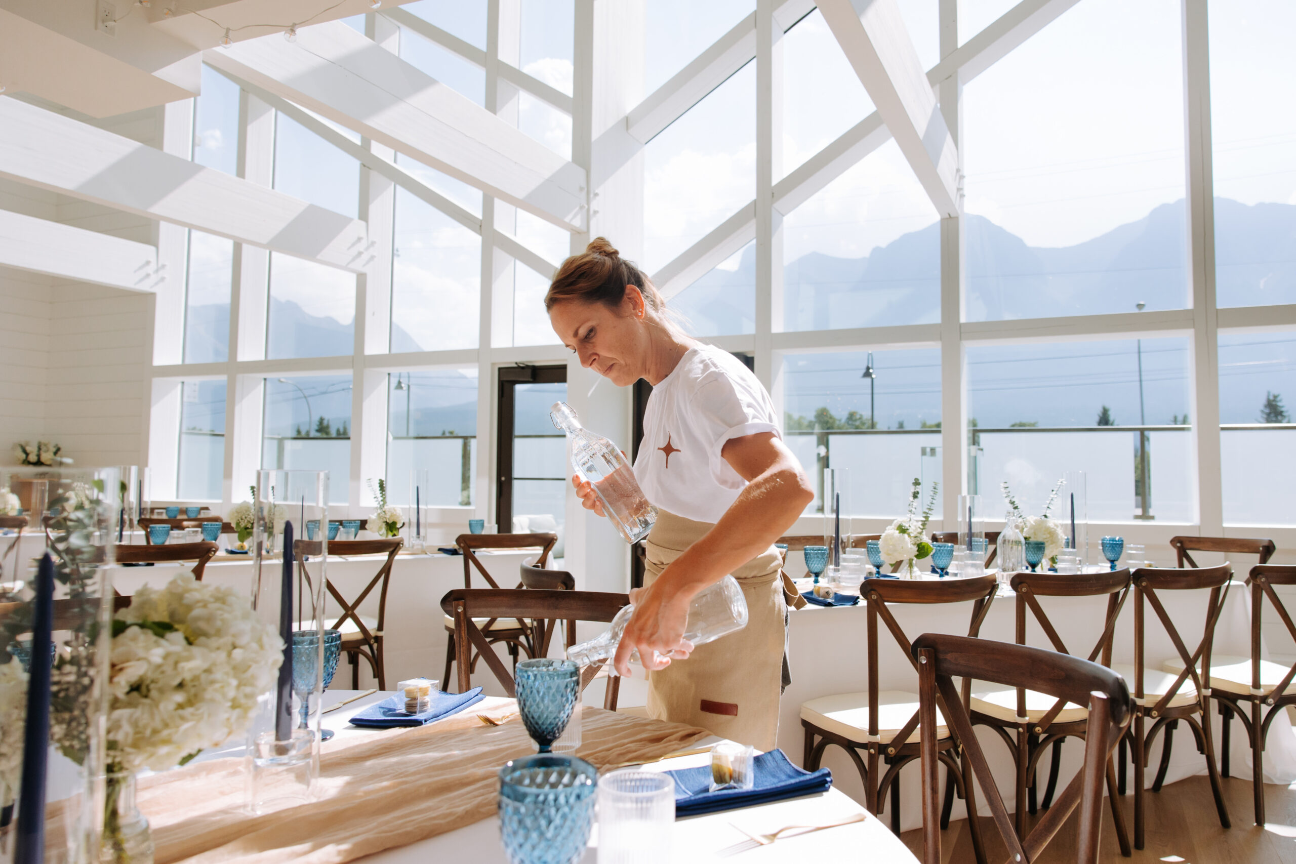 A staff member at The Gem carefully fills glasses with water while setting up a wedding reception with blue accents and mountain views.