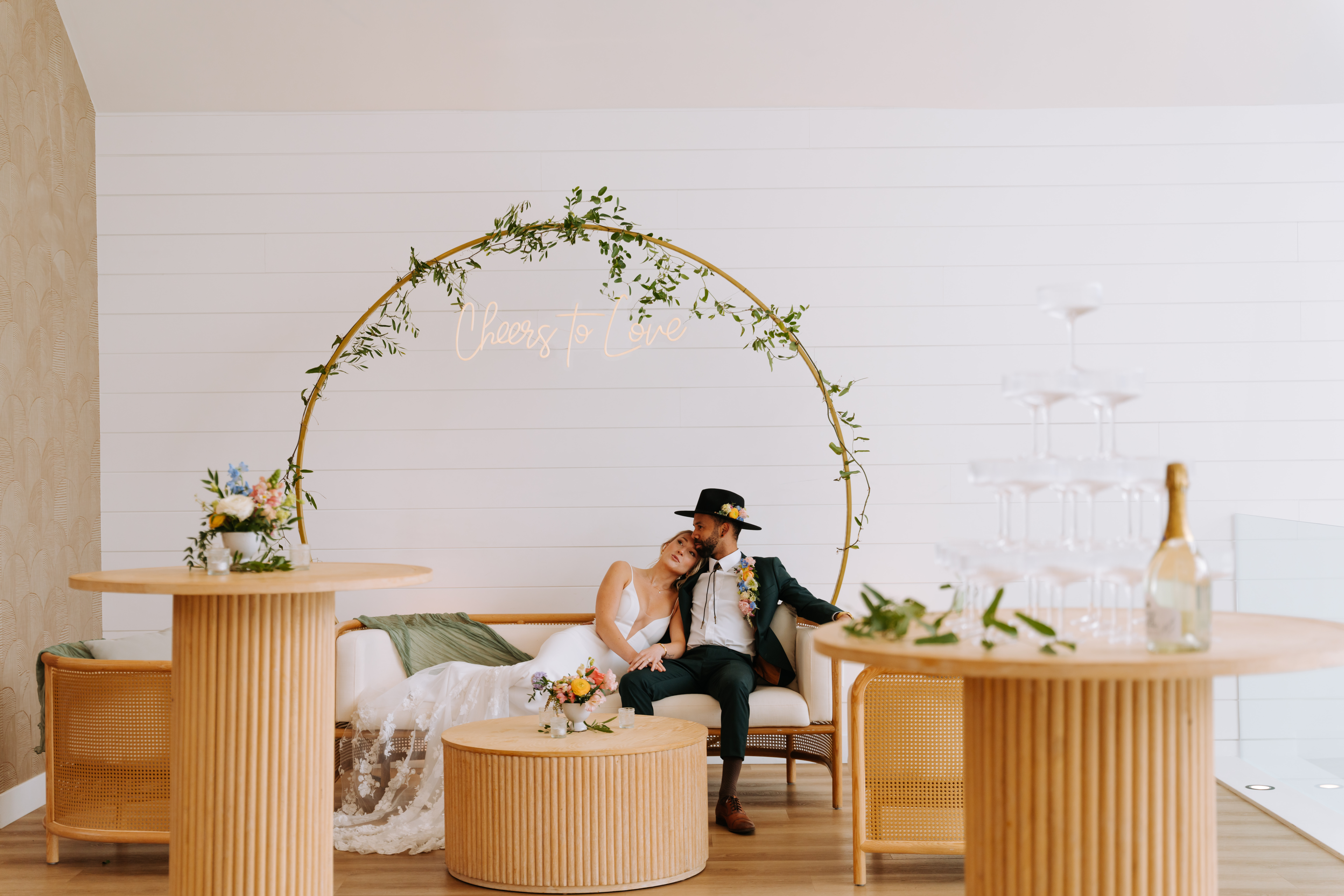 Bride and groom sitting in a modern wedding lounge at The Gem in Canmore, beneath a neon sign reading "Cheers to Love."