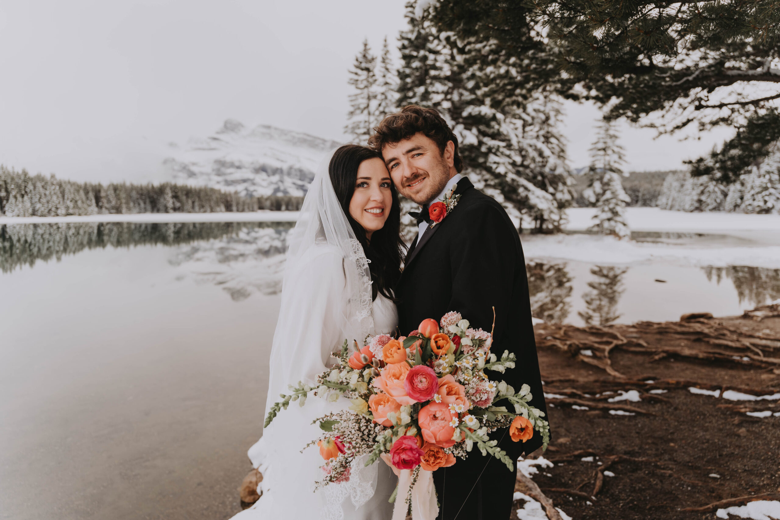 Bride and groom posing by a snowy lakefront at Lake Minnewanka with a vibrant bouquet and mountain reflections in Banff National Park.